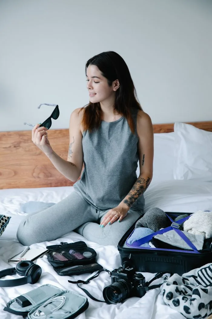 Young woman sitting on bed packing suitcase for a trip, holding sunglasses.